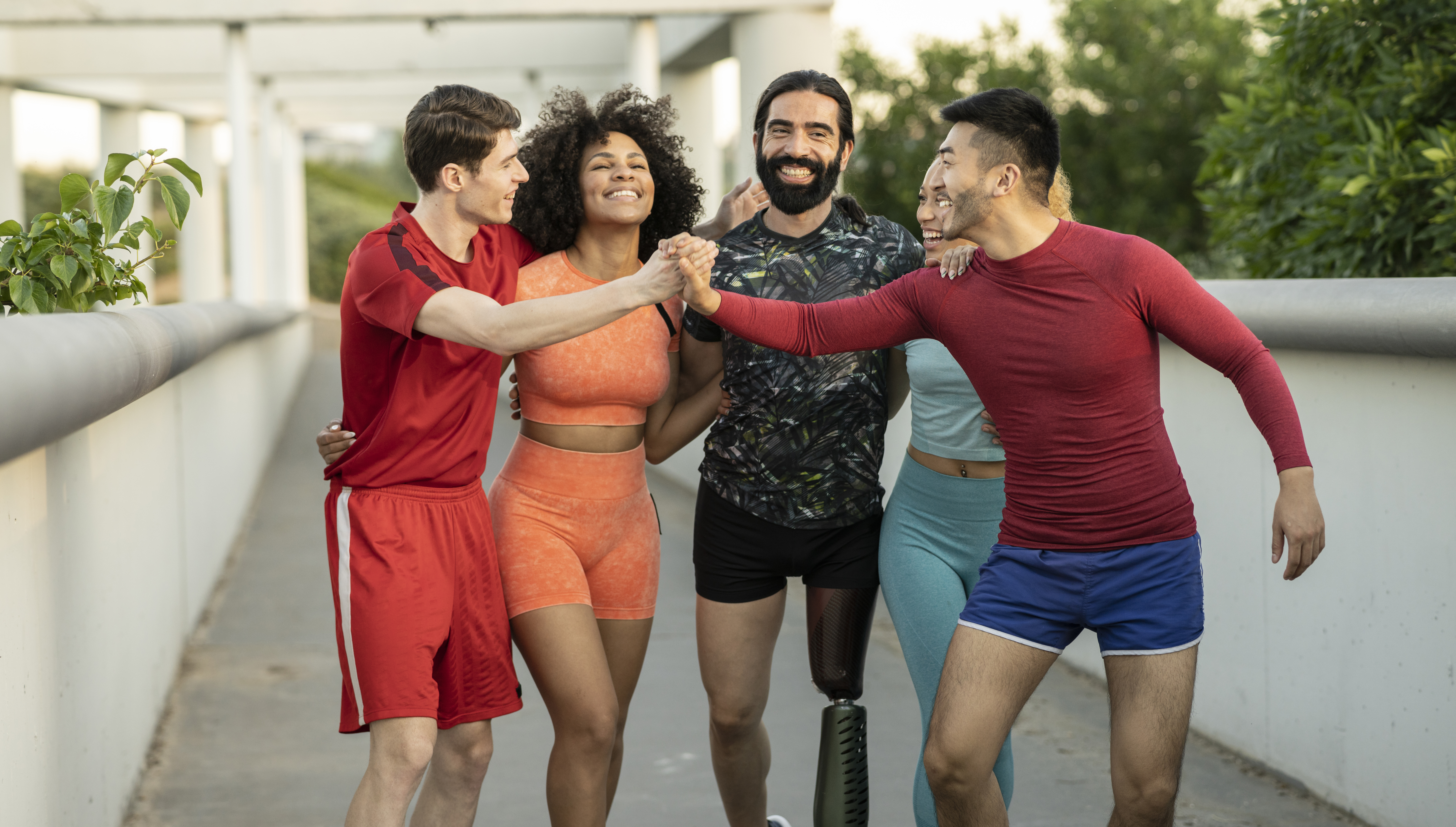 'Diverse group of five friends in workout gear celebrating with a fist bump on an outdoor path, one man with a below-the-knee running blade prosthetic, STEPtember fitness challenge