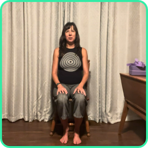 Video thumbnail for a 15 minute seated yoga class, woman sitting on a chair with hands resting on her knees in front of curtains.